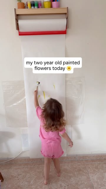 A toddler painting yellow flowers on a paper roll on the wall